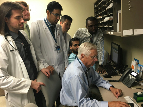 Residents standing behind a professor while he is sitting in front of a computer